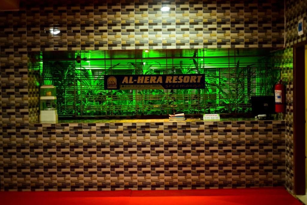 Empty reception desk at Al Hera Resort with backlit green-lit indoor plant display and an illuminated 'AL-HERA RESORT Sreemangal' signboard