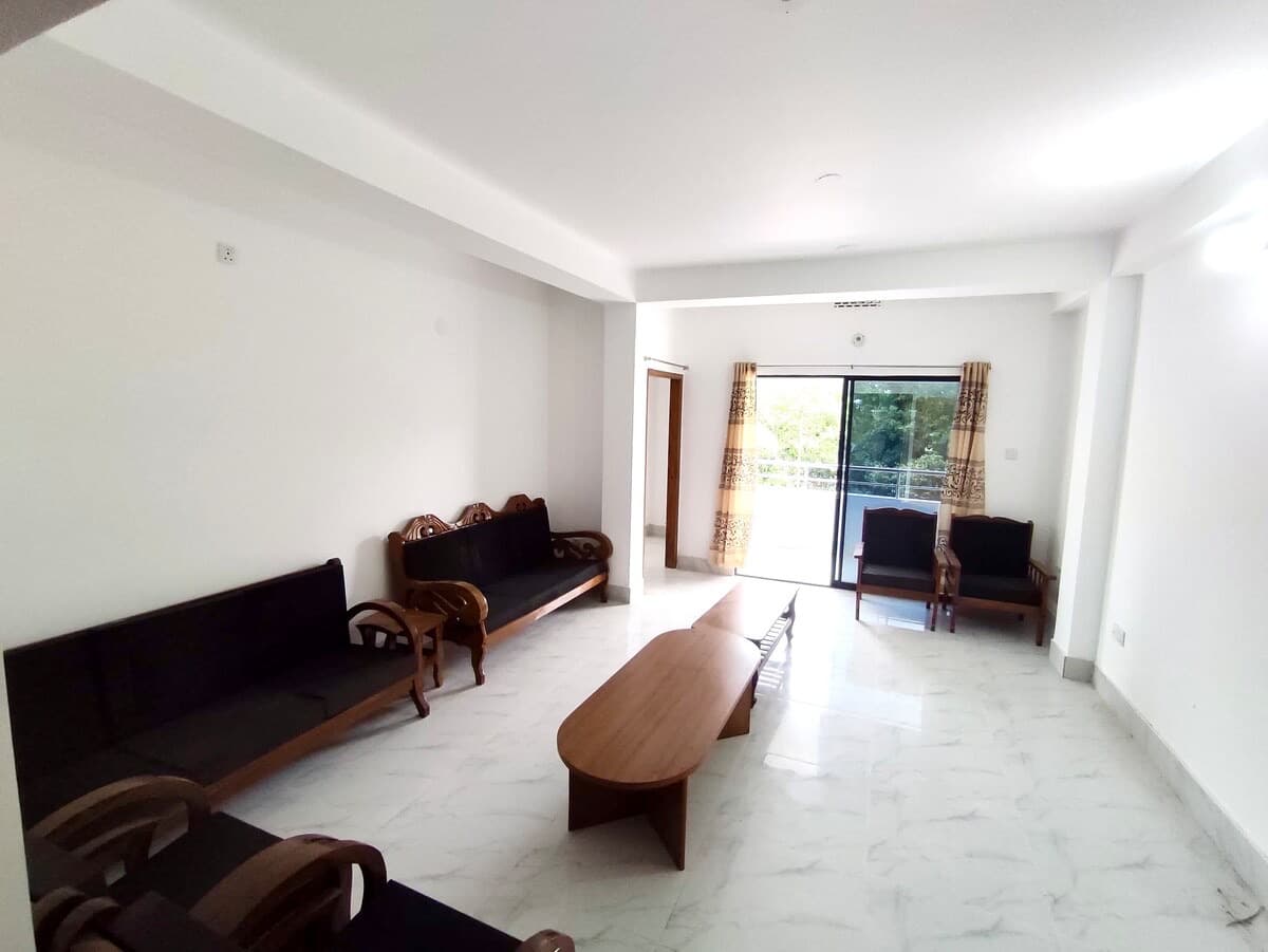 Living room with two long wooden-framed black sofas, an oval wooden coffee table, two armchairs, and a sliding glass door opening onto a balcony with greenery outside