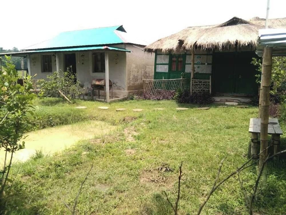 Grounds view of Bondhur Bari Eco Resort showing a tin-roofed cottage and a thatched-roof green-doored cottage set among a grassy lawn