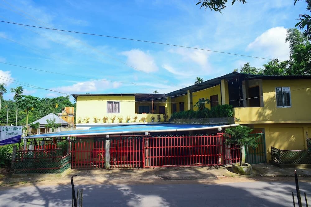 Front view of the yellow two-storey guesthouse building with the red gate and blue pool-cover awning, with a signboard reading 'Brishti Bilash Guest House' visible at the property entrance