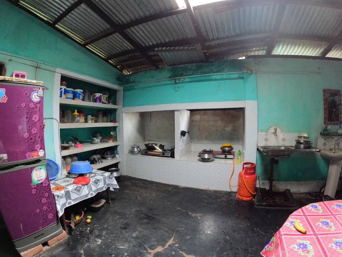 Household kitchen under a corrugated tin roof, with a purple Marcel refrigerator, open shelves of cookware, twin gas burners on a raised tiled platform, an LPG cylinder and a steel sink against teal walls