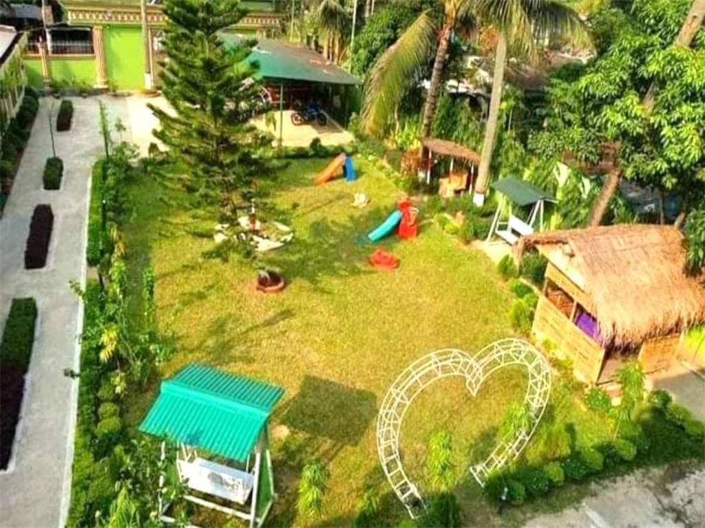 Aerial daytime view of the resort's lawn playground — slide, swing, a thatched-roof play hut, and a white heart-shaped arch, bordered by a paved walkway