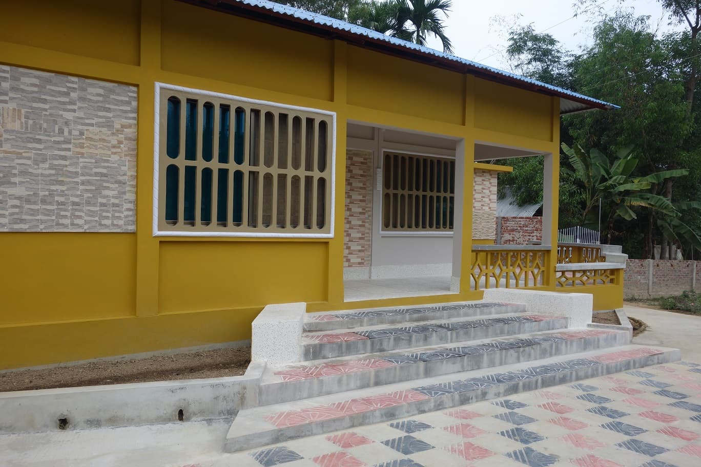 Newly built single-storey guest block painted mustard yellow with a blue corrugated-tin roof, a covered veranda behind ornate yellow concrete balustrades, stacked stone-tile wall panels and moulded concrete window grilles, fronted by pink-and-grey stamped-concrete paving