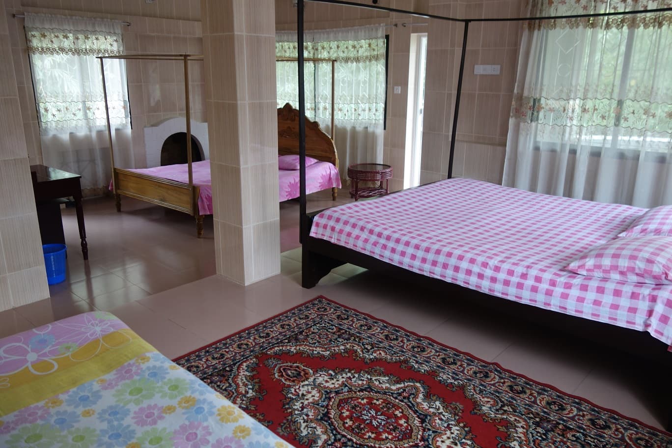 Family room with several four-poster beds under metal mosquito-net frames — pink checked, pink floral and pastel floral bedspreads — stone-tile clad walls, lace-trimmed net curtains, a writing desk and an ornate red patterned rug on a tiled floor
