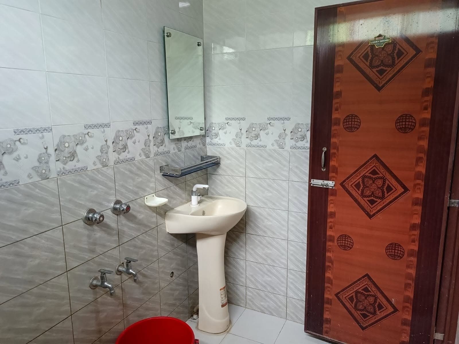 Tiled bathroom with a pedestal wash basin, frameless wall mirror, glass shelf, floral tile border, low bucket-fill taps and a wood-pattern door with a sliding bolt