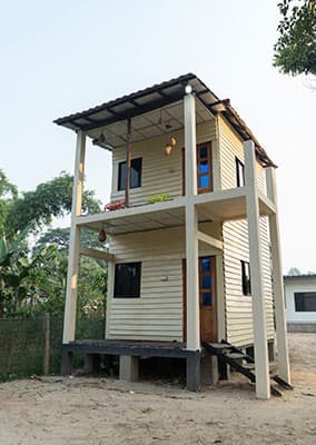 Two-story cream-painted wooden guest cottage on stilts with a corrugated brown roof, covered upstairs veranda, and an external staircase, surrounded by trees