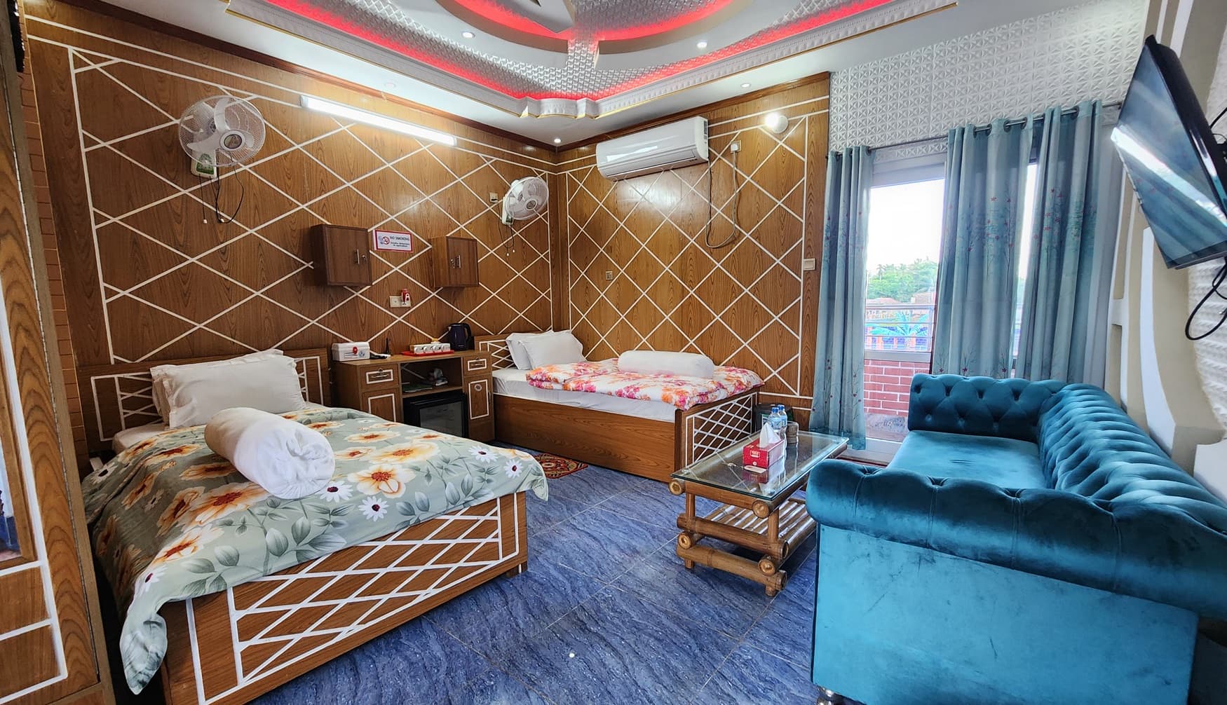 Twin-bed room at Mukta Nagar Resort with wood-panel walls, red accent ceiling lighting, a teal tufted sofa, and a glass-and-bamboo coffee table