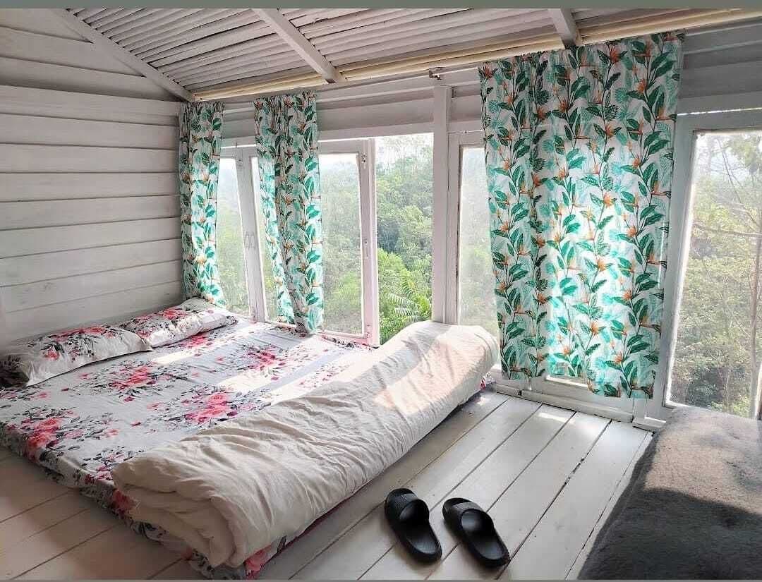 Loft bedroom with a floor mattress dressed in floral bedding, tropical-print curtains, and floor-to-ceiling windows overlooking the forest