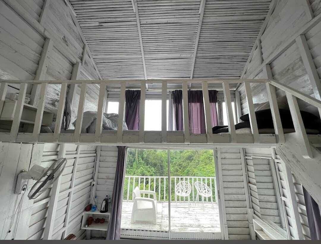 Upward view inside the wooden cottage showing the sleeping loft, ladder railings, and glass doors opening to the balcony and forest below