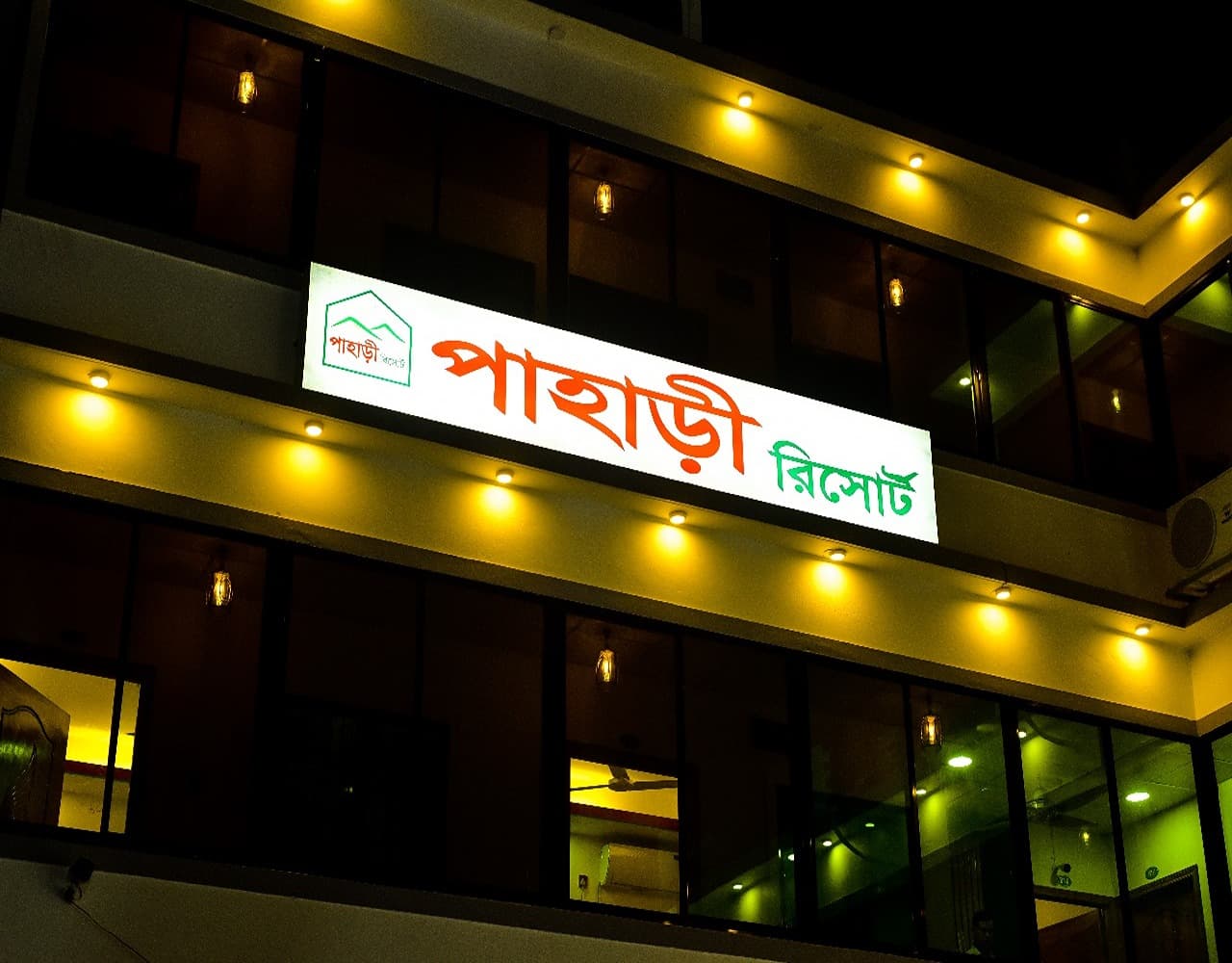 Close-up of the illuminated signboard reading "Pahari Resort" in Bangla, mounted on the building facade at night