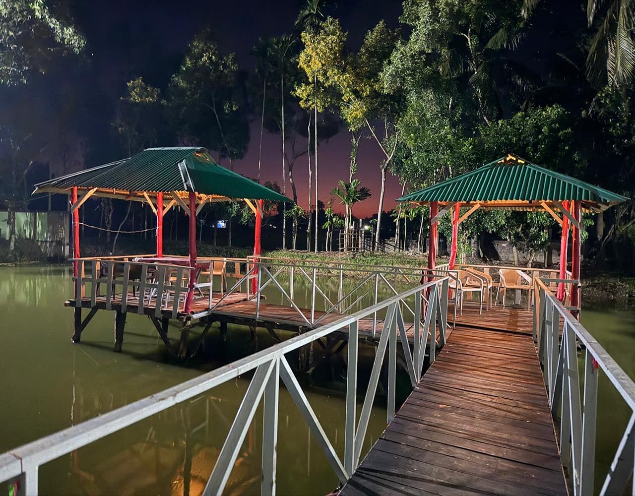 Twin wooden gazebos with green roofs connected by a railed timber walkway over a pond on the property at dusk