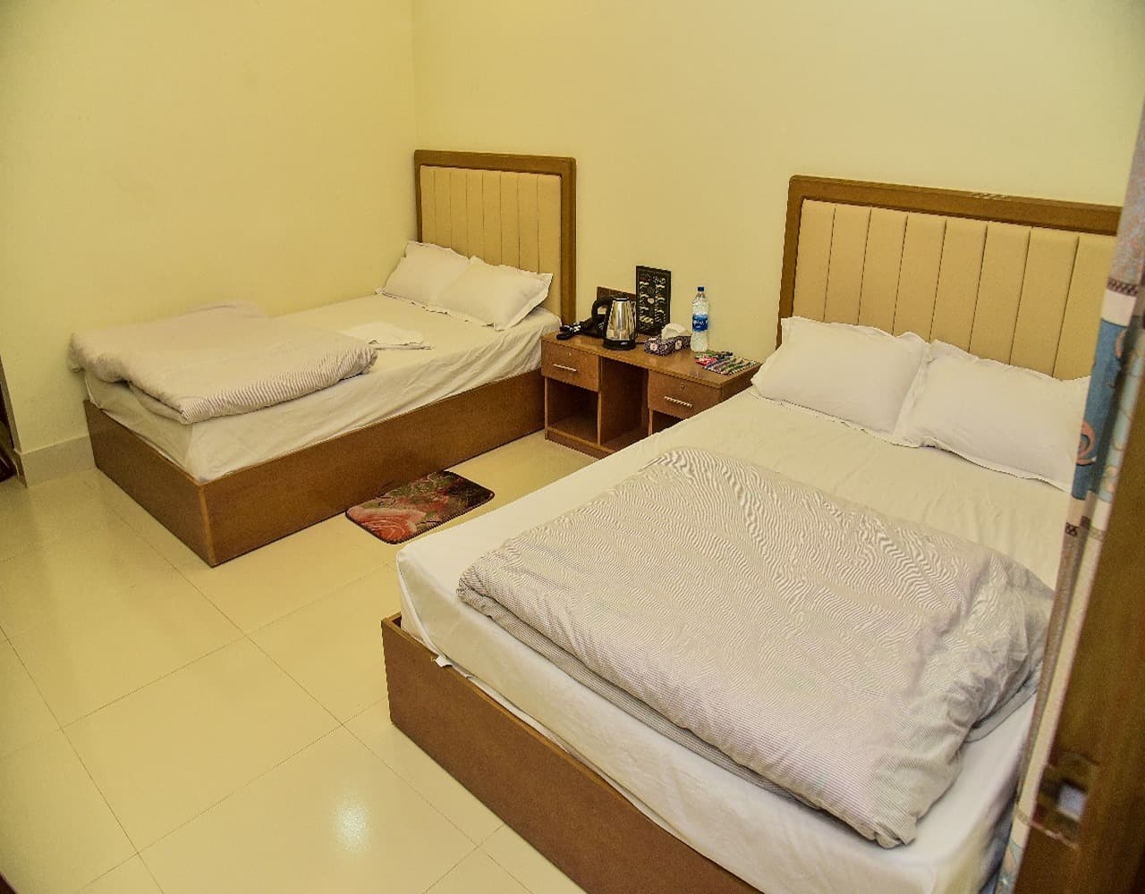 Twin-bed and queen-bed room with cream leatherette headboards, a shared nightstand with kettle and amenities, and a rose-patterned floor mat