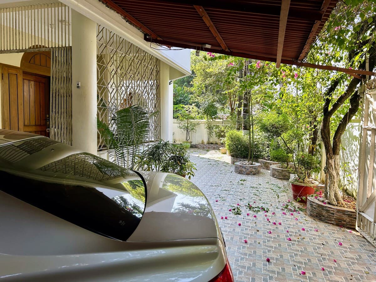 Covered driveway with a parked car and lush garden courtyard beside the private room near Sreemangal railway station