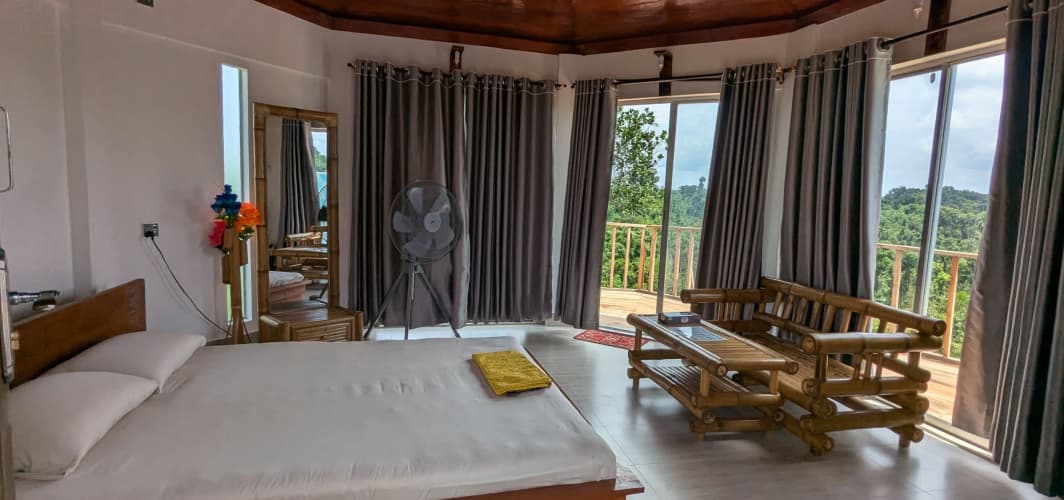 Bedroom interior with curved wooden ceiling, a bed with white linens, standing fan, bamboo sofa and coffee table, and floor-to-ceiling windows opening onto the deck