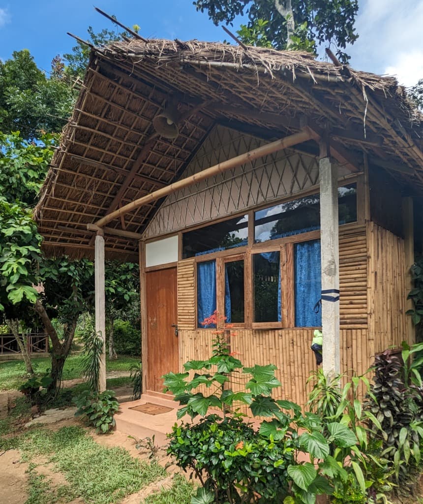 A thatched bamboo cottage at Shanti Bari with a wooden door, diamond-lattice gable and blue-curtained windows