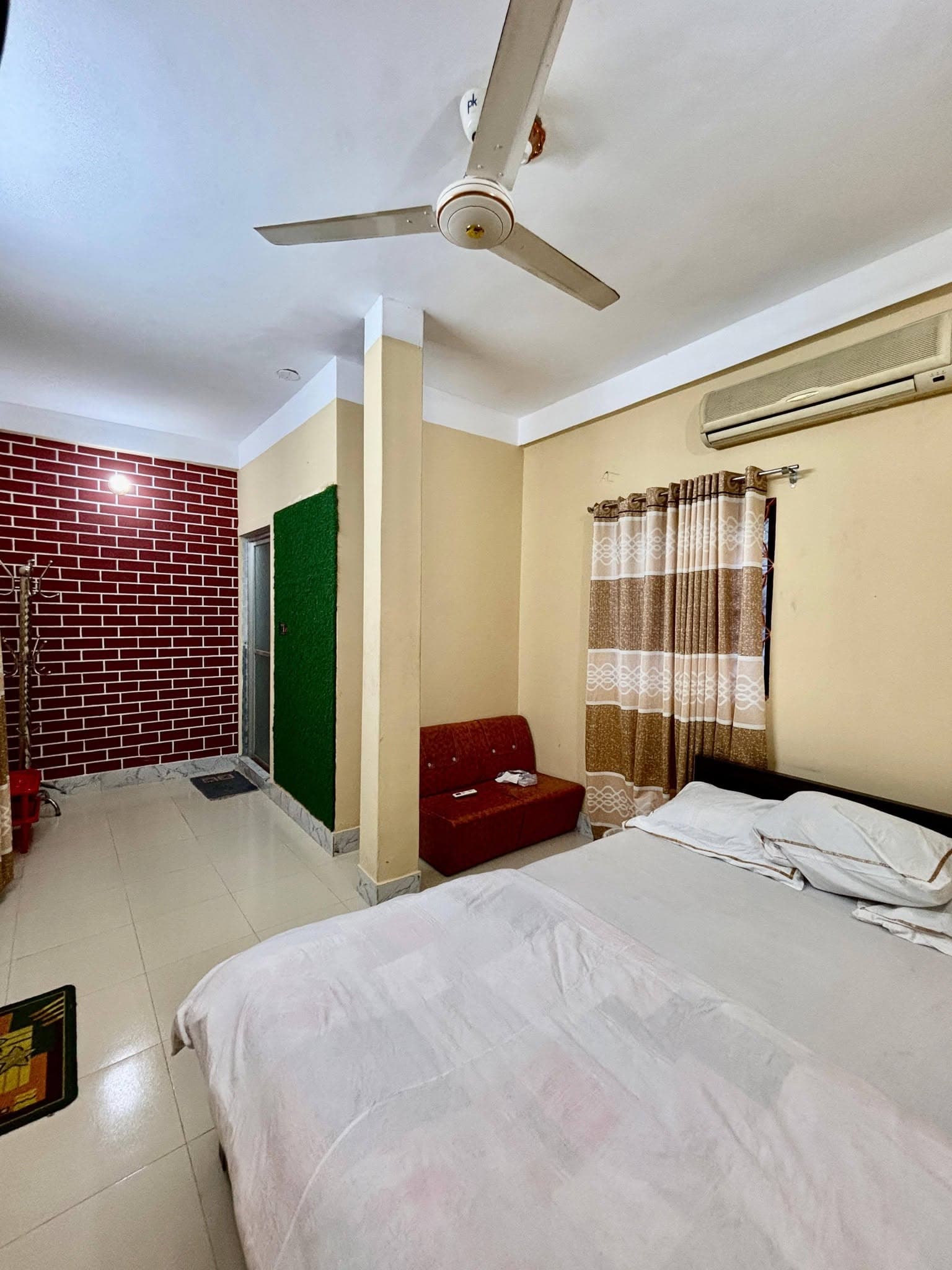 Guest room with a double bed, wall-mounted air conditioner, red loveseat, striped curtains, and a red-brick accent wall visible through an open doorway