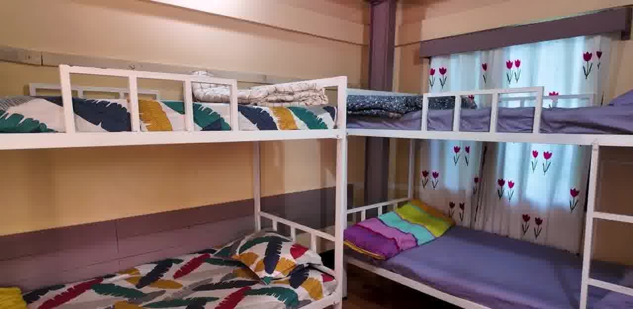 Dorm-style guest room with two white metal bunk beds, the lower bunks made up in colourful feather-print and folded pastel bedding, against peach walls with a lilac dado and tulip-print curtains at the window