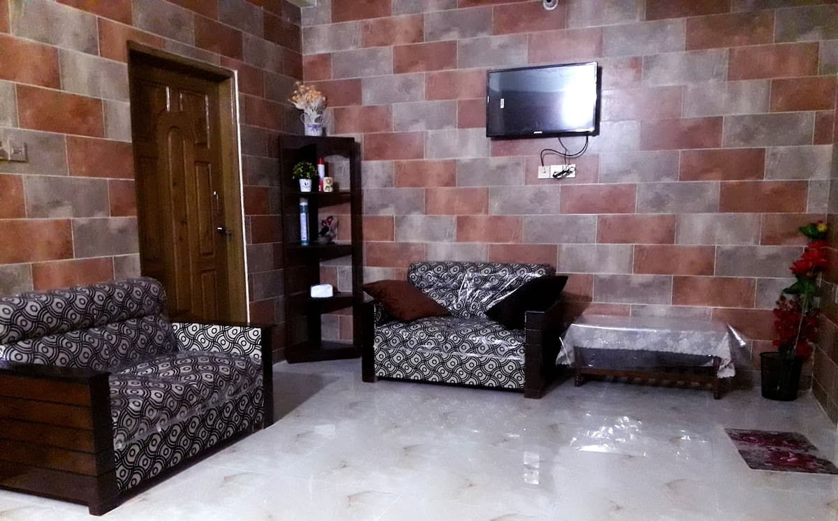 Sitting room at Sreemangal Cottage with two black-and-white patterned sofa sets still part-wrapped in plastic, a dark wood corner display shelf, a wall-mounted flat-screen TV on a brick-effect tiled feature wall, and a low wooden table on glossy cream marble-look flooring