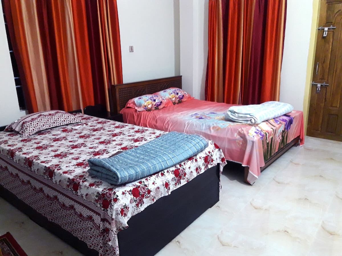 Twin-bed guest room at Sreemangal Cottage — one bed under a red-rose floral bedspread, the other under pink floral linen, each with a folded blue quilt, against white walls hung with floor-to-ceiling orange and red curtains and a grilled window at the left