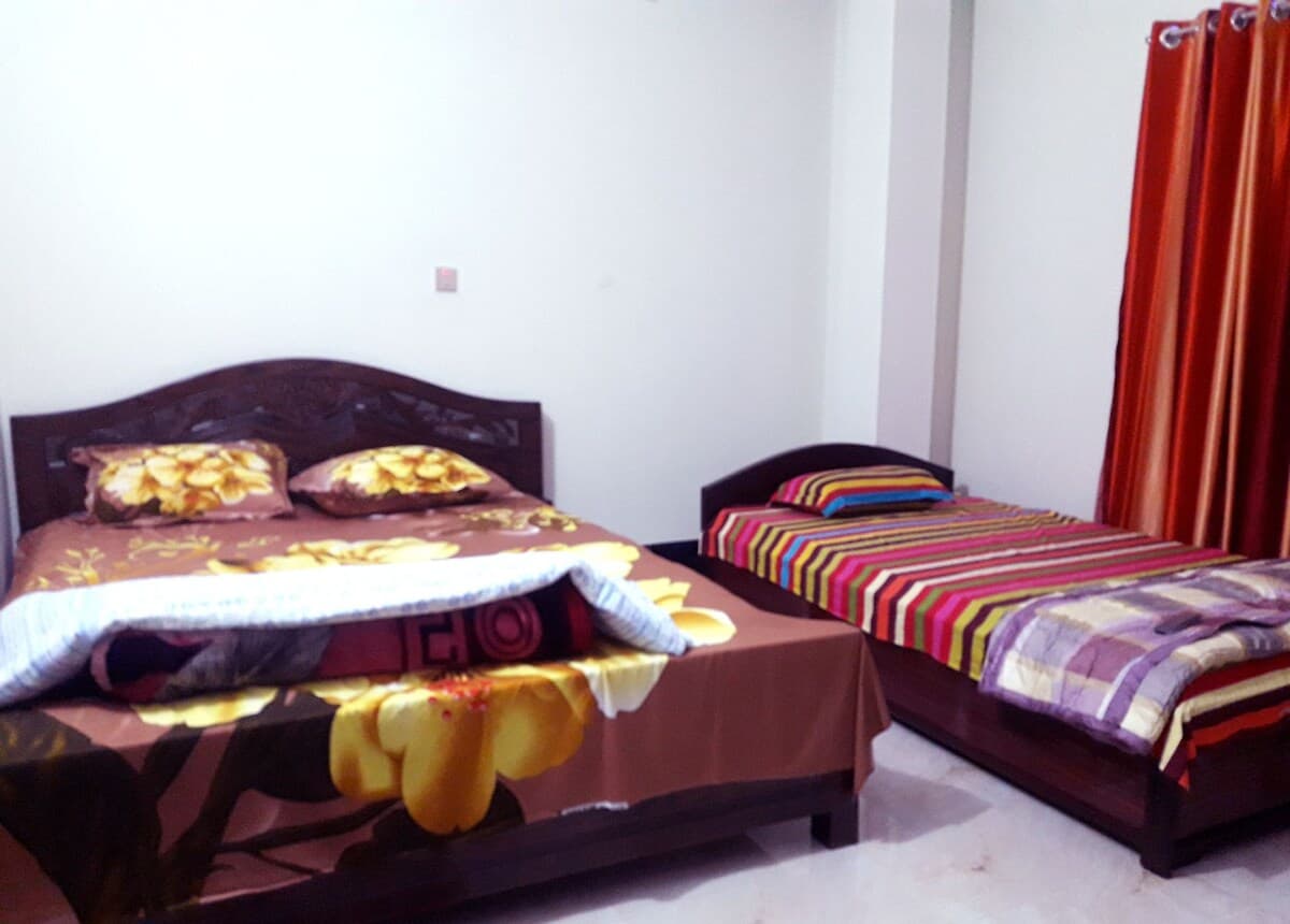 Guest room at Sreemangal Cottage with a carved dark-wood double bed dressed in brown and yellow floral linen beside a separate single bed under a multicoloured striped cover, red-orange curtains at the right and pale walls