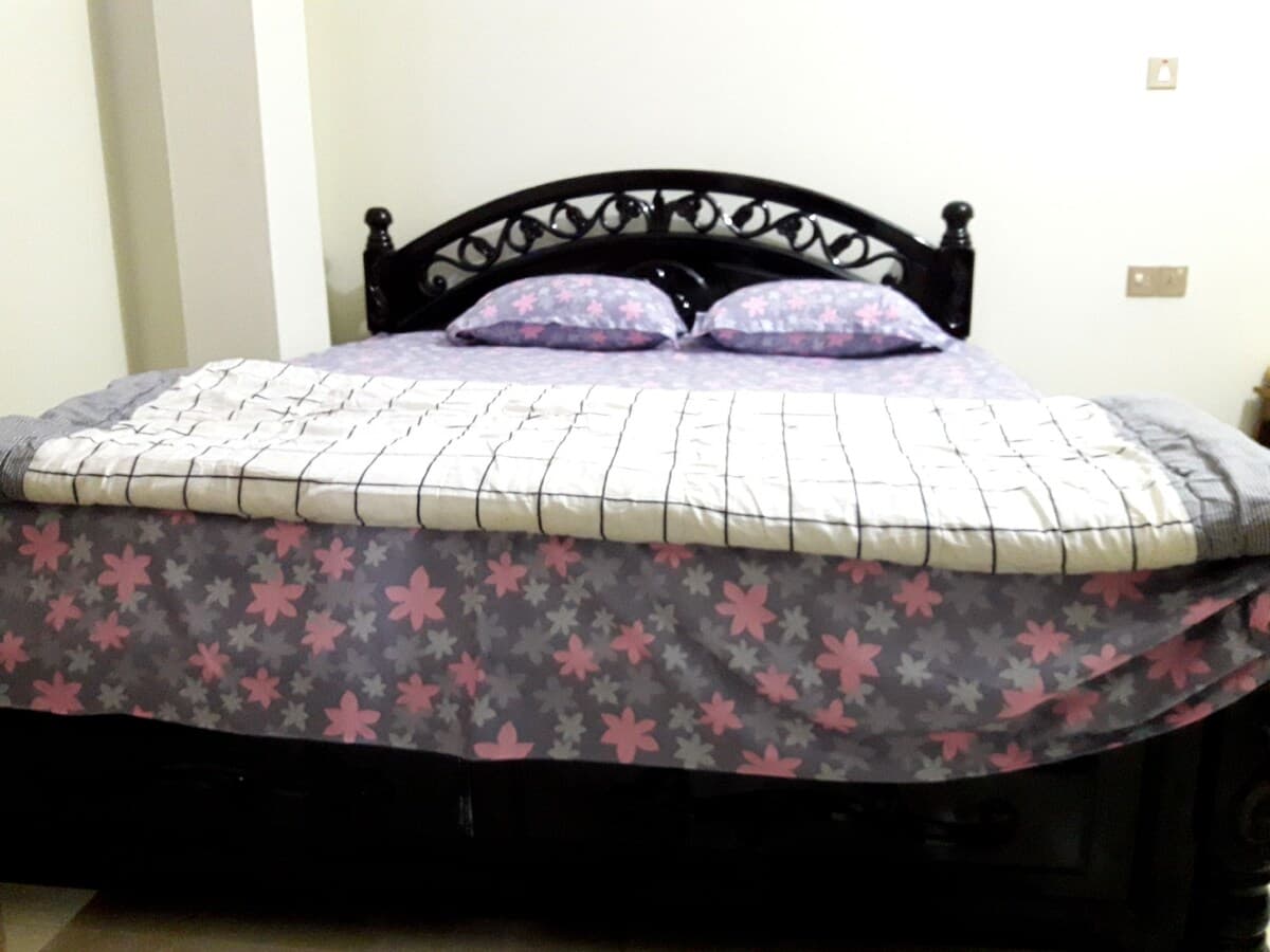 Double bed at Sreemangal Cottage with an ornate black wrought-iron-style headboard, two pink-flowered pillows, a grey and pink floral valance and a white quilted throw with a black grid pattern