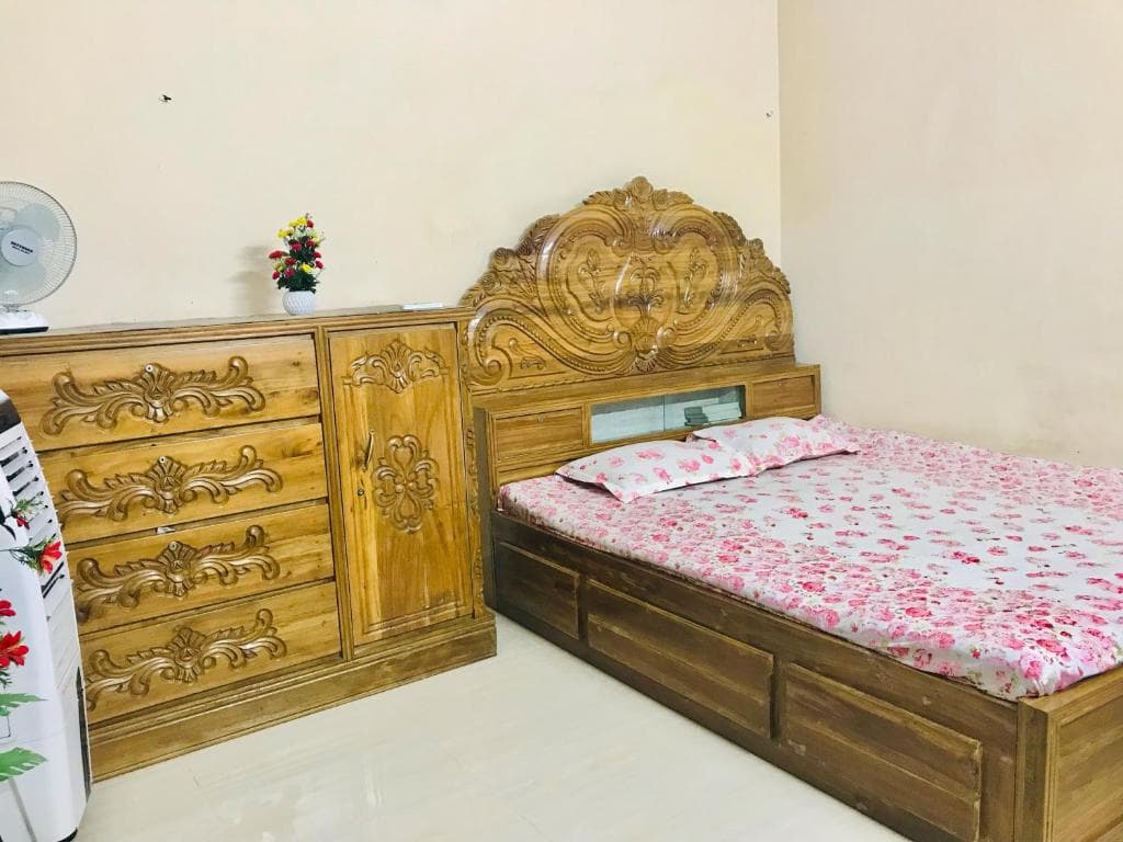 Bedroom with an ornately carved teak bed and matching carved chest of drawers and cabinet, pink floral bedspread and pillow, a small vase of artificial flowers, a pedestal fan and a portable evaporative air cooler on a pale tiled floor