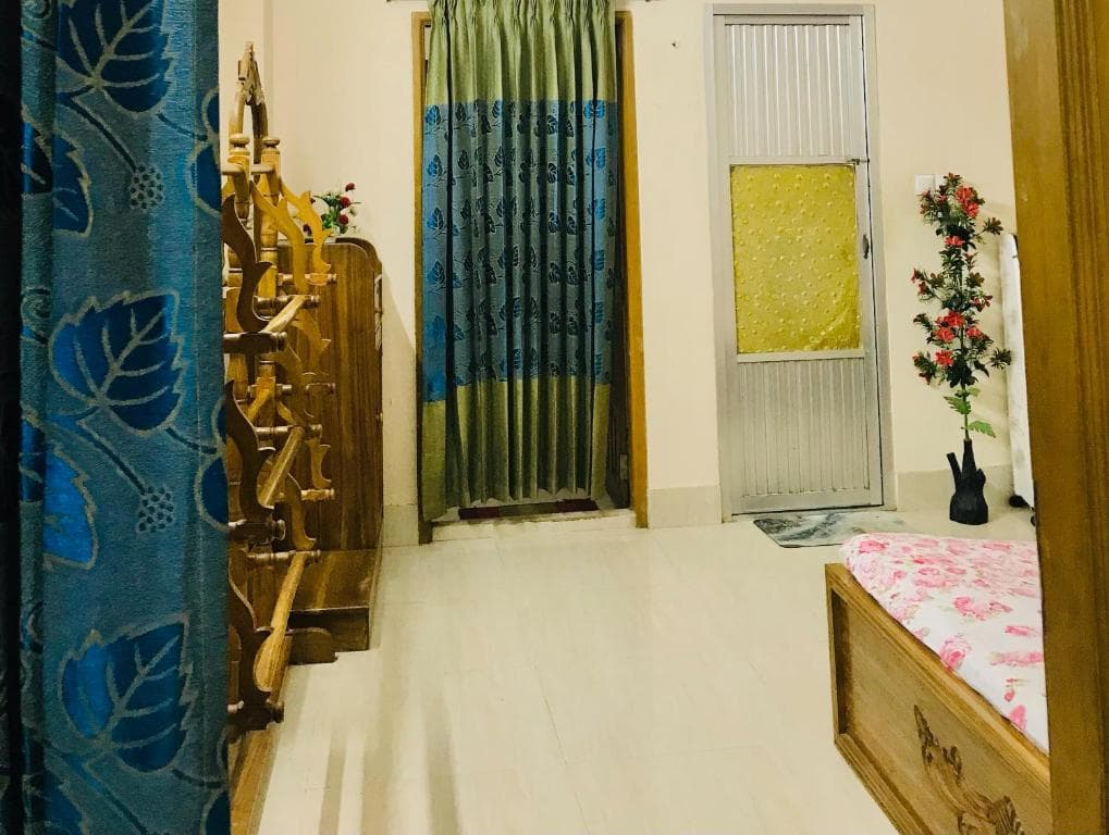 Corner of a bedroom looking toward the entrance: a blue leaf-print curtain in the foreground, an ornately carved wooden bed headboard along the left wall, a green-and-blue curtained doorway, a ridged aluminium door with a yellow frosted panel, an artificial red-flowered plant, and the corner of a floral-covered bed at right