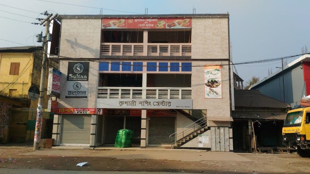 The full three-storey Sultan Restaurant & Guest House building seen from Railway Station Road, with its black 'Sultan Restaurant & Guest House' signboard on the first floor, a red Bangla restaurant banner across the top floor and the Rupali Shopping Center shutters at street level