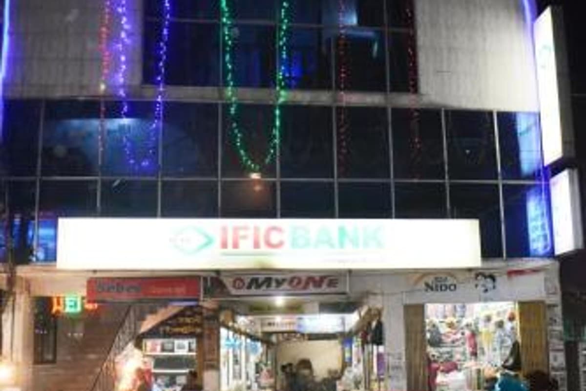 Night view of the Razzaque Tower building entrance in Sreemangal, with the ground-floor IFIC Bank branch and adjoining shops lit up, string lights strung across the upper floors