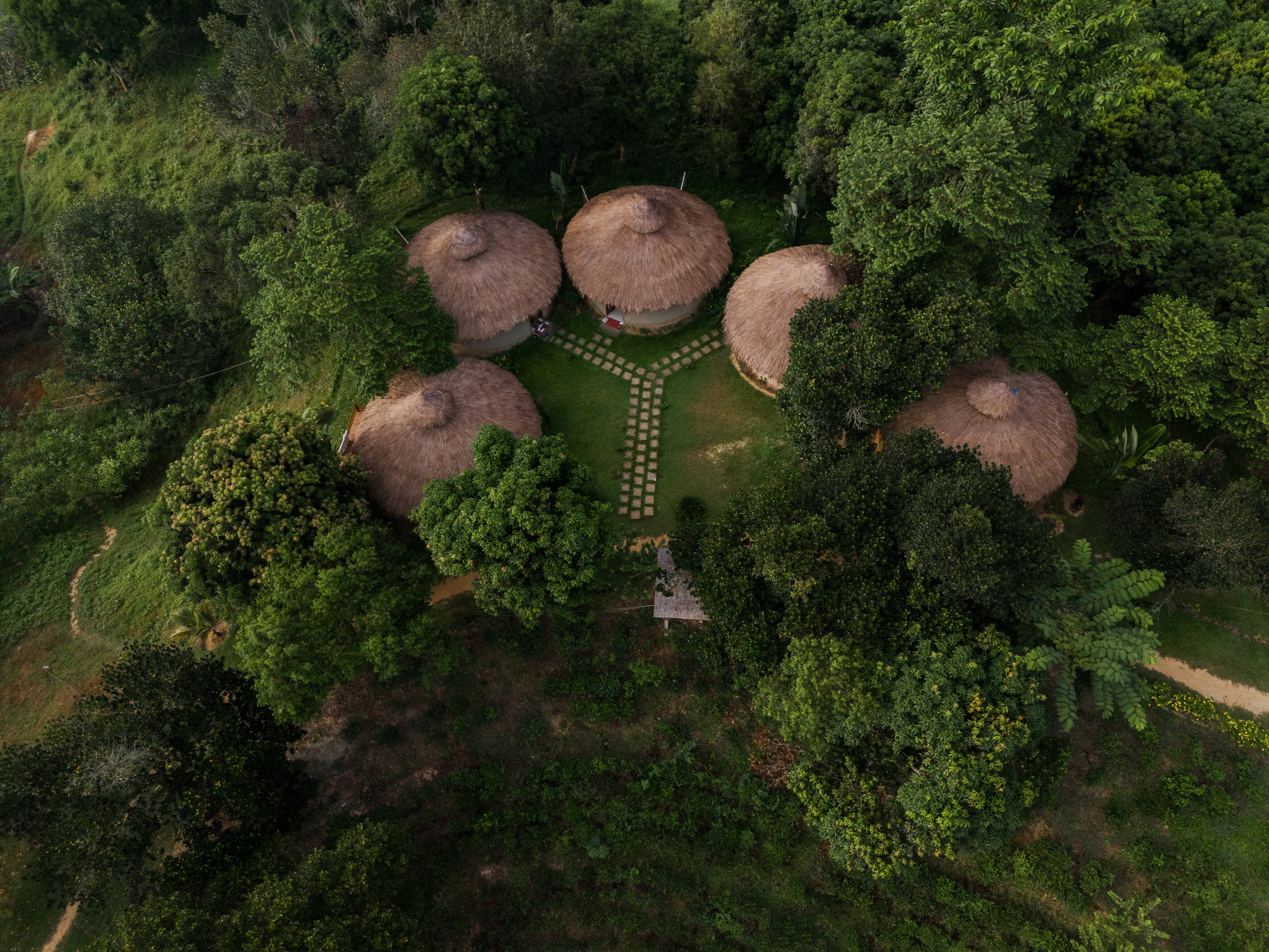 A drone view of five round thatched huts arranged around a stone-paved courtyard, ringed by forest