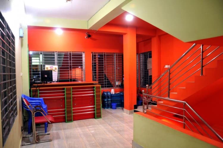Orange-and-green reception lobby with a wooden front desk, blue plastic chairs, and a staircase to the right