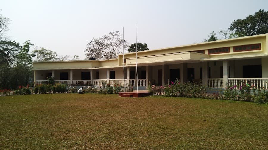 Full frontal view across the lawn of the long single-storey cream guest house, with a continuous columned veranda, cream turned-baluster railing, a row of identical guest room doorways, a raised block at right carrying a band of red-framed clerestory windows, two flagpoles on a plinth, and flower beds along the veranda edge