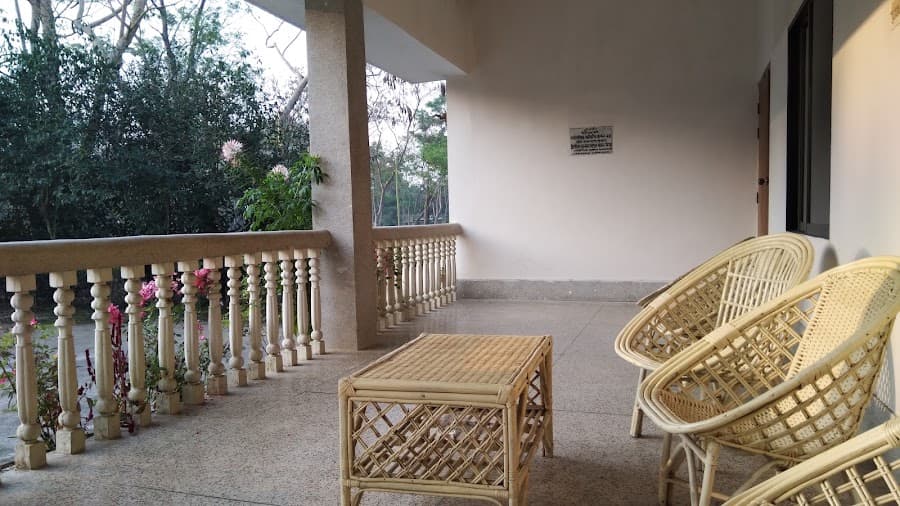 Seating corner on the guest house veranda with two pale wicker tub chairs and a wicker coffee table on a polished terrazzo floor, a terrazzo-clad square column and cream turned-baluster railing looking out onto flowering shrubs, and a small framed Bangla notice plaque on the wall beside a dark doorway