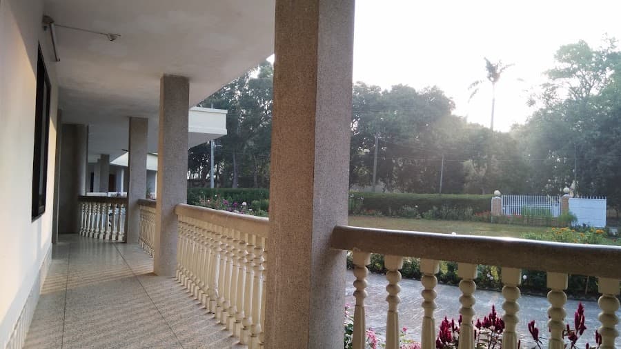 View along the guest house veranda at dusk, terrazzo-clad square columns receding down the covered walkway with a cream turned-baluster railing on the right, overlooking the lawn, flower borders and clipped hedge, with the compound's white perimeter fence and brick gate pillars in the far distance